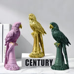 Three exotic parrot figurines in pink, gold, and dark green on cylindrical perch stands, marble surface