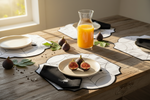 A styled dining table scene with a glass carafe of orange juice, figs, plates, and black napkins on a rustic wooden table in warm natural light.