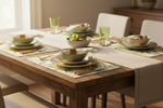 A styled dining table setting with layered ceramic plates, light green bowls, matching green drinking glasses, and leaf-patterned placemats on a wooden table with cream chairs.