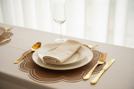 A styled table setting with white plates, a folded cream napkin, gold-toned cutlery, and a clear wine glass on a beige tablecloth in front of a softly blurred window.