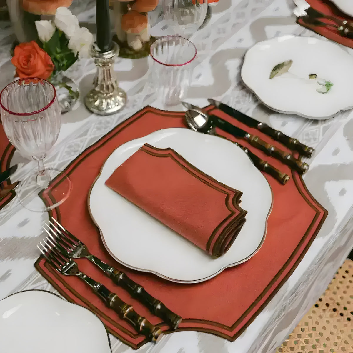 Rustic Red Placemats on dining table, terracotta-red square mat with notched corners and dark border, white plate, red napkin, bamboo cutlery