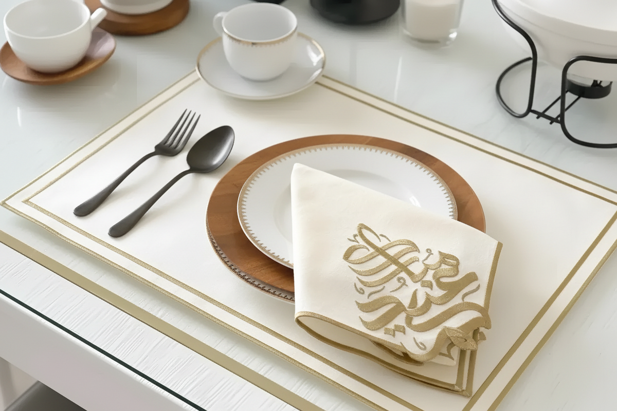 A styled table setting featuring a cream-colored rectangular placemat with gold borders, a white ceramic plate with a gold-rimmed edge, a folded cream napkin with gold Arabic calligraphy, silver cutlery, and white teacups on a white table.
