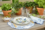 A styled outdoor table setting featuring a white ceramic dinner plate with a colorful bird and floral illustration, a matching white napkin with blue floral detailing, a scalloped white placemat with blue trim, and clear glass goblets. The setting is decorated with potted green plants in terracotta pots on a light wooden table.
