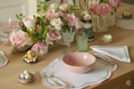 A styled dining table setting with a pink ceramic bowl, scalloped white placemat with pink and green trim, silver cutlery, pink and green glassware, and a floral centerpiece with pink and white flowers. A small bunny figurine and Easter-themed decor are visible in the background on a light wood table.