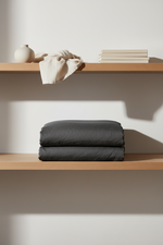 A folded dark charcoal gray bedding set, likely a duvet cover and pillowcases, is neatly stacked on a wooden shelf. Above it, a light beige linen cloth and a small ceramic vase sit on another shelf against a warm off-white wall, creating a minimalist, modern interior scene.