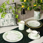 White ceramic dinnerware set with green lily of the valley botanical print, styled on dark green velvet table runner with lemon tree backdrop