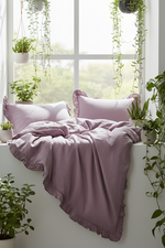 A bed dressed in dusty lavender bedding with ruffled edges and matching pillows, styled in a bright room by a window with hanging green plants.