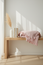 A soft blush pink fringed throw blanket is draped over a light wood bench in a minimalist room with warm sunlight streaming across the wall and floor. A white vase with pampas grass and three books sit beside the blanket.