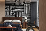 A modern bedroom interior featuring a bold black and white geometric patterned accent wall behind a bed with white pillows and a navy throw blanket. A dark gray chair, a small table with a lamp, and a window with blinds are visible in the room.