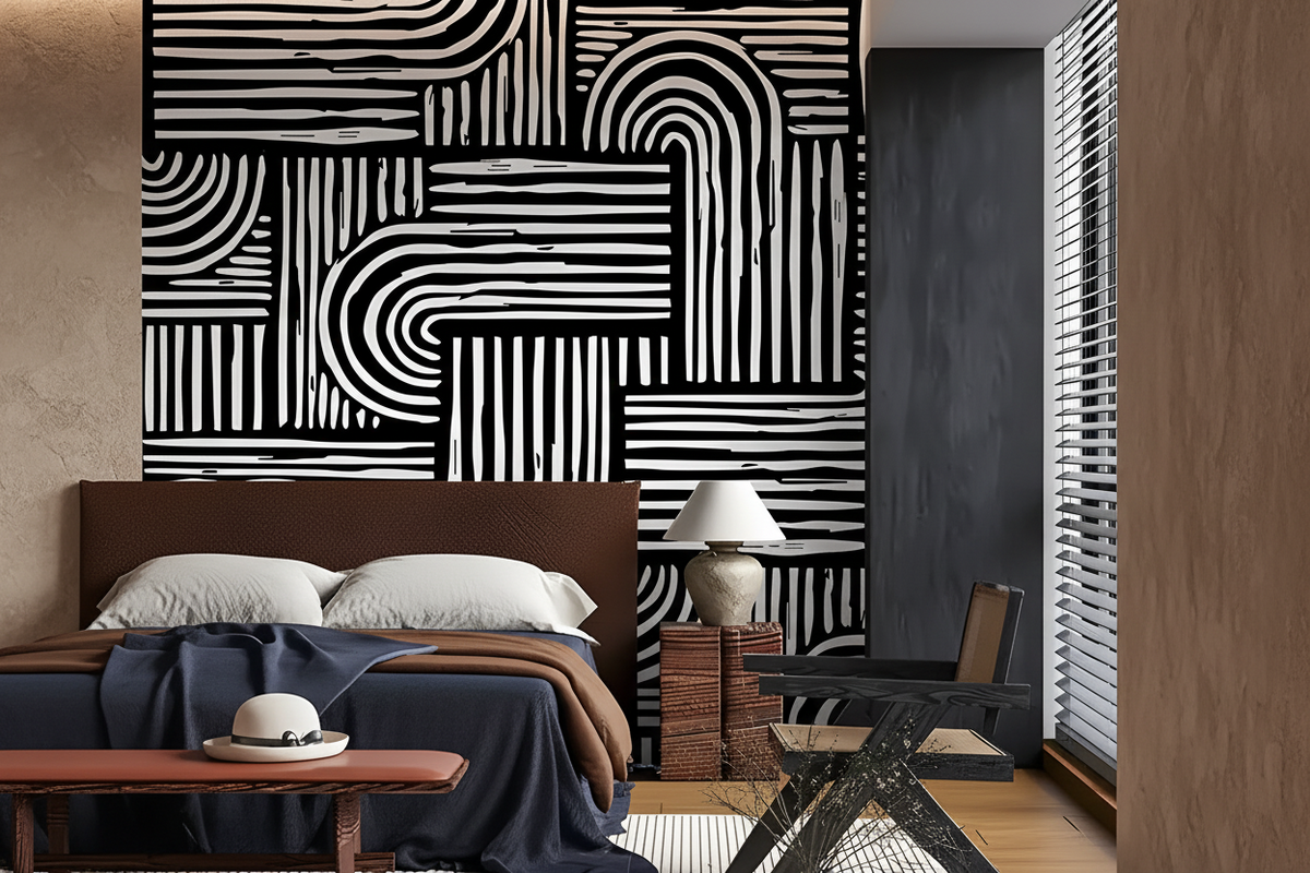 A modern bedroom interior featuring a bold black and white geometric patterned accent wall behind a bed with white pillows and a navy throw blanket. A dark gray chair, a small table with a lamp, and a window with blinds are visible in the room.