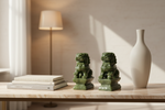 Two green stone lion statues on a light marble console table in a softly lit room, with stacked books, a tall white vase, a floor lamp, and warm sunlight casting shadows.