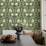 Room interior with floral-patterned wallpaper, table with bread and vase, and pillows on a bench.