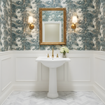 Bathroom with pedestal sink, gold-framed mirror, and floral arrangement against decorative wallpaper.