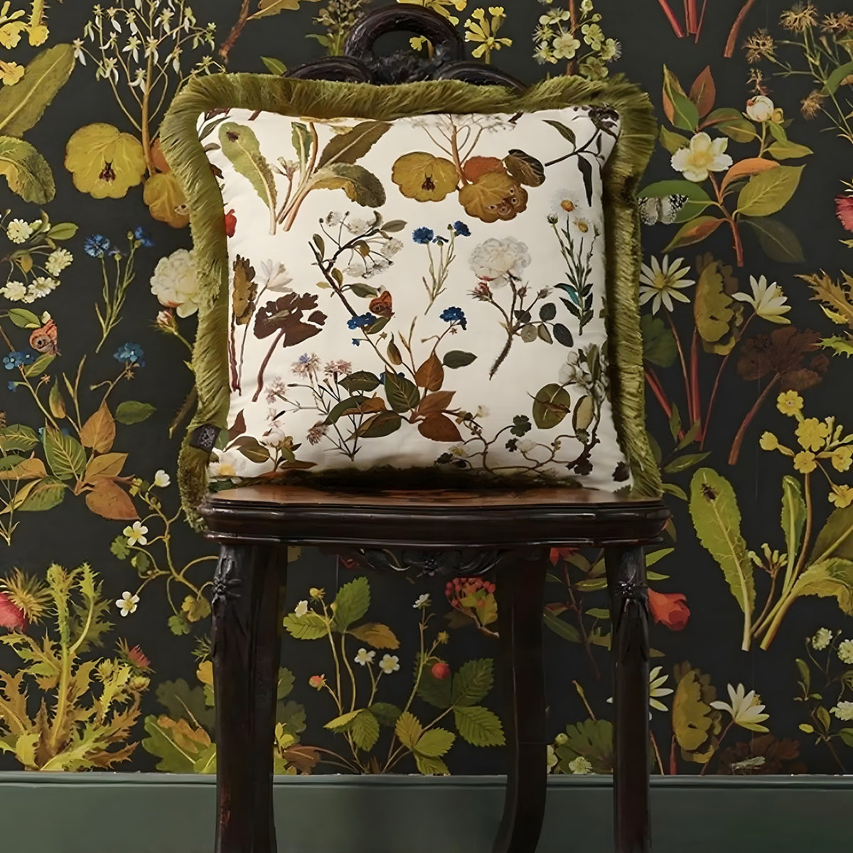 Decorative pillow with floral pattern on a wooden chair against a floral wallpapered wall.