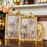 Two decorative gold photo frames with animal heads on a wooden surface, with flowers and a candle in the background.