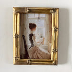 Framed artwork of a woman sitting by a window with a gold frame.
