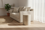 A modern beige upholstered sofa with matching throw pillows in a bright, minimalist living room, alongside a cylindrical patterned side table and a potted green plant.