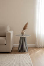 A modern side table with a black and white vertically striped tapered base and a round white top, styled in a minimalist living room next to a beige armchair with a small vase of pampas grass on top.
