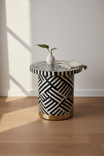 A round side table with a black and white geometric bone-inlay pattern and a gold metal base, styled with a small white vase and a folded napkin on a light wood floor against a white wall.