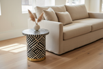 A modern side table with a black and white geometric inlay pattern and a gold base, styled next to a beige fabric sofa in a bright living room.