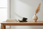 A styled interior scene showing a light wood console table against a white wall, with stacked neutral books, a dark snail figurine, and a ribbed cream vase holding dried pampas grass.