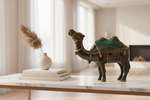 A decorative camel figurine with a dark brown body and a teal-green and gold patterned saddle, displayed on a white marble-topped table in a bright, modern living room setting.