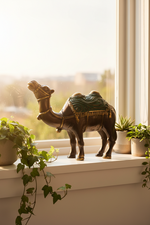 A decorative camel figurine with a dark brown body and green-and-gold saddle, displayed on a windowsill with potted plants in a softly lit room.