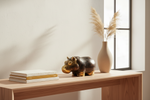 A decorative hippo figurine in dark brown and gold sits on a light wood console table next to a beige ceramic vase with dried pampas grass, with stacked books in the foreground against a warm off-white wall.