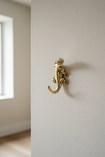 A polished brass gecko-shaped wall hook mounted on a textured off-white wall, with a softly blurred interior room and window in the background.
