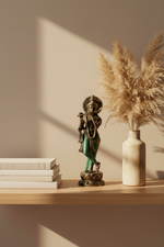 A decorative bronze-toned statue of a flute-playing deity stands on a light wood shelf beside a cream ceramic vase filled with dried pampas grass, with stacked books on the left. Warm sunlight casts shadows on a beige wall.