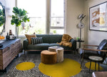 A stylish living room featuring an abstract maze golden sun hand-tufted wool area rug. The rug's bold design complements a modern setting with a green sofa, chic furniture, and large windows. Ideal for enhancing living room and dining room spaces, this large area rug adds a touch of elegance to formal dining rooms.