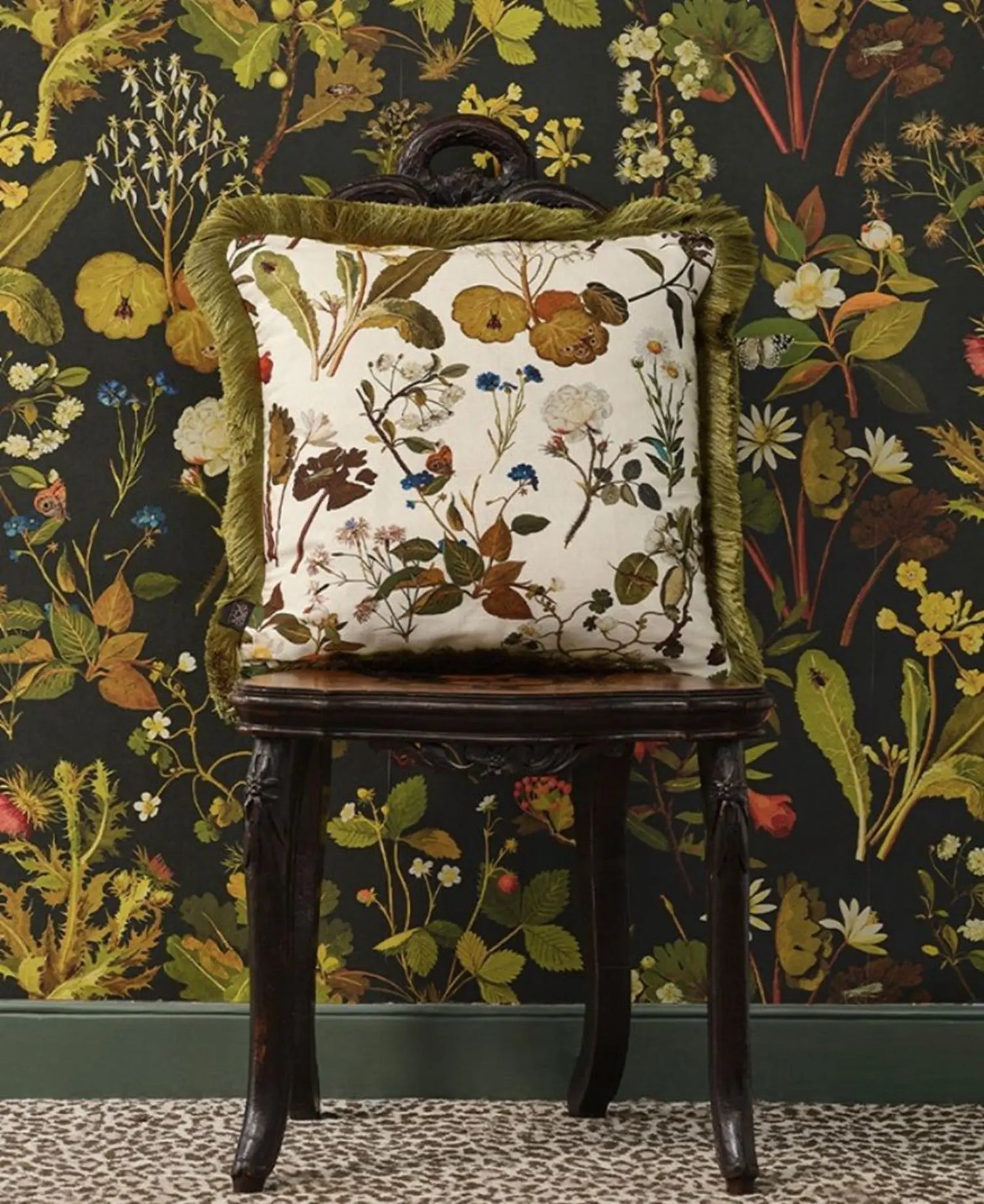 Decorative pillow with floral pattern on a wooden chair against a floral wallpapered wall.