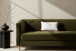 An olive green upholstered sofa in a styled interior, with a white accent pillow featuring a fox illustration and red pom-pom corners, next to a black side table with a black vase and dried branches.