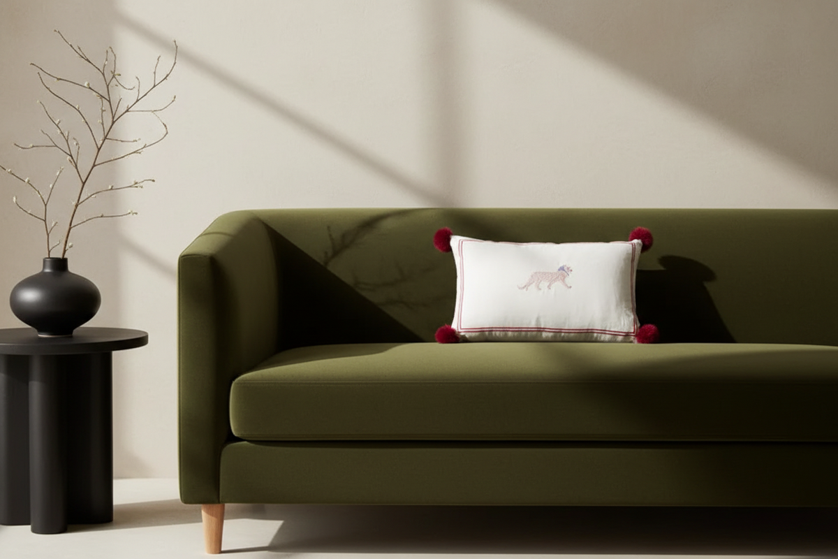 An olive green upholstered sofa in a styled interior, with a white accent pillow featuring a fox illustration and red pom-pom corners, next to a black side table with a black vase and dried branches.