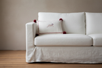 A white slipcovered sofa with a white accent pillow featuring a small embroidered dog and red pom-poms, set against a beige wall and light wood floor.