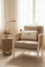 A beige upholstered armchair with woven cane sides and a decorative throw pillow featuring a pink embroidered animal, styled in a bright, sunlit room next to a carved wooden side table with flowers and a floor lamp.