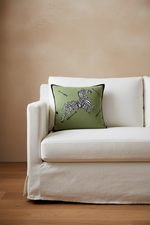 A square olive green throw pillow with a black and white zebra print and the word 'SABAJA' embroidered on it, resting on a white slipcovered sofa against a warm beige wall.