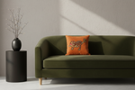 An olive green velvet sofa in a modern living room setting, featuring a bright orange throw pillow with a embroidered tiger design. A black cylindrical side table with a black vase and dried branches stands to the left against a textured light gray wall with soft diagonal sunlight shadows.