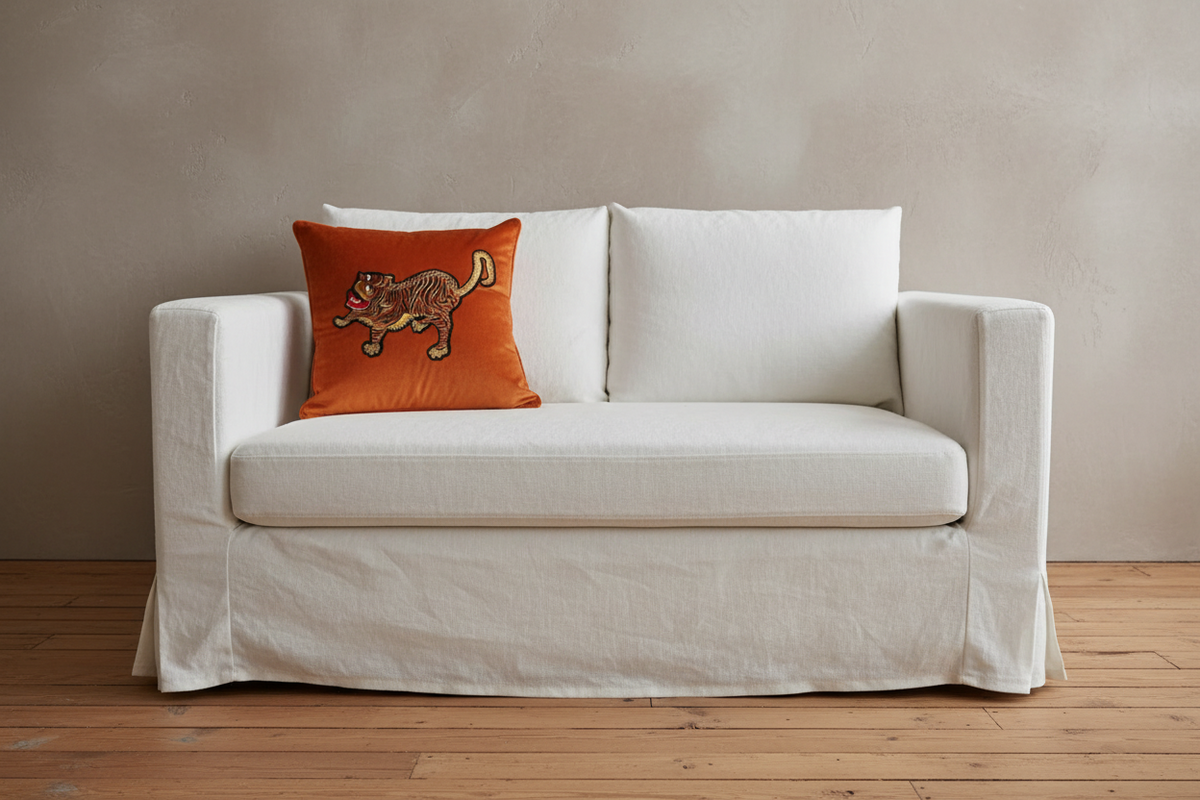 A white slipcovered loveseat or sofa with two back cushions and a single orange accent pillow featuring a embroidered cat design, set against a textured beige wall and light wood floor.