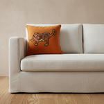 An olive green velvet sofa in a modern living room setting, featuring a bright orange throw pillow with a embroidered tiger design. A black cylindrical side table with a black vase and dried branches stands to the left against a textured light gray wall with soft diagonal sunlight shadows.