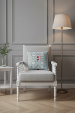 An olive green velvet sofa in a modern living room setting, featuring a square light blue throw pillow with a delicate pink and red floral pattern, next to a small black side table with dried branches.