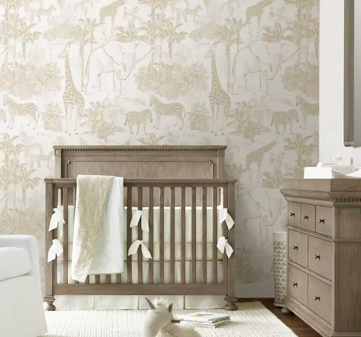 A styled nursery interior featuring a wooden crib with white bedding and tassels, a matching wooden dresser, and a wall covered in safari-themed wallpaper with giraffes, zebras, and palm trees.