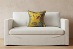 A square decorative throw pillow with a vibrant yellow background and a detailed botanical and bird pattern, styled on a cream-colored fabric sofa against a textured beige wall.