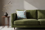 An olive green velvet sofa in a styled room setting, featuring a decorative light blue throw pillow with a white bird and red floral pattern, next to a small wooden side table with a dark vase of dried flowers.