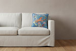 A square decorative throw pillow with a colorful floral and horse design, resting on a light beige fabric sofa against a textured beige wall and light wood floor.