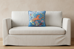 A cream-colored slipcovered sofa with a square blue throw pillow featuring a colorful embroidered horse and floral pattern, set against a textured beige wall and light wood floor.
