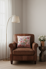 A brown leather armchair in a softly lit room, styled with a square floral throw pillow. A tall floor lamp stands to the left, and a small side table with white flowers is to the right.