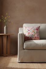 A beige upholstered armchair in a styled room setting, featuring a square pink floral throw pillow. A small wooden side table with a gray vase of yellow flowers stands to the left against a textured warm brown wall and light wood flooring.