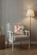 A light gray upholstered armchair with a white spindle-style frame sits in a room with wood-paneled walls and a lit floor lamp. A square throw pillow with a colorful butterfly print rests on the chair.