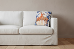 A square decorative throw pillow featuring an illustration of two giraffes, styled on an olive green velvet sofa in a modern living room setting.